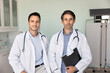 © fizkes - Two confident male doctor colleagues in white uniform coats with stethoscopes posing together in clinic examination room, looking at camera, holding pad with medical records. Practitioners portrait