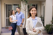 © fizkes - Happy female realtor posing outdoors with rental accommodation and couple of renters in background, looking at camera, smiling, holding digital tablet. Successful real estate agent woman portrait