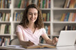 © fizkes - Preparing for exam. Smiling confident teen female student sit at desk in library study online use e-learning platform on notebook do academic assignment get information in books web take notes by hand