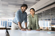 © fizkes - Two cheerful Indian startup colleagues meeting for teamwork, posing for corporate portrait, leaning on office table with paper marketing reports, looking at camera, smiling