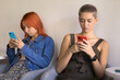 © fizkes - Digital addiction. Two diverse generation-z females teenagers spend time indoors wait in line sit in cozy armchairs keep silence use mobile phones for texting chatting online surfing social networks