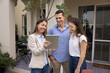 © fizkes - Happy real estate agent woman showing content on tablet to young couple of tenants, renters, house buyers, standing at apartment entrance outside, talking, laughing, discussing rent or purchase terms