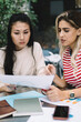 © BullRun - Puzzled Asian woman analyzing paper report during collaborative brainstorming with Spanish freelance colleague discussing solutions for business projecting, diverse students preparing to exams