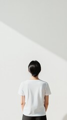 Poster - Woman with black hair wearing white t shirt standing in front of white wall with light from window creating diagonal shadow, perfect for mockups