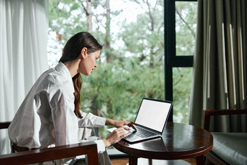 Canvas Print - Relaxed young woman working on a laptop at home, wearing a casual white shirt in a serene, green outdoor setting with soft natural light