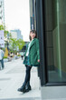 © ultramansk - A cheerful young woman outdoors in a vibrant urban setting, dressed in a green coat, with blurred golden lights and greenery in the background, capturing a lively and candid moment.