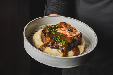 Wall Mural - A chef holds a plate featuring a succulent piece of meat with rich gravy, served on a bed of creamy mashed potatoes, garnished with fresh herbs. Perfect for gourmet food showcases.