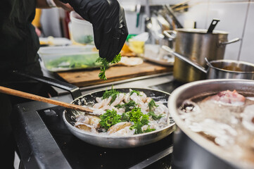 Canvas Print - A chef's hand adds fresh parsley to a pan of seafood, enhancing the dish's flavor. Busy kitchen environment, with pots and fresh ingredients in the background. Perfect for culinary themes.