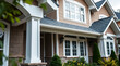 © MatandSterre - Exterior view of a two-story tan-brown house with white trim, featuring multiple windows, a covered porch with white columns, and landscaping elements including shrubs and flowers.