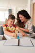 © Dorde - Caucasian mother in striped shirt assists elementary-age son with homework assignment at home study space, demonstrating educational support and guidance.