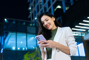 Portrait of young Asian businesswoman walking in city street at Night. Attractive Smiling Asian woman using a mobile phone standing in street and looking around on street of modern city at night.