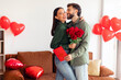© Home-stock - Happy couple celebrating Valentine's Day, man giving flowers and gift card to beloved woman, posing in living room with love and warmth