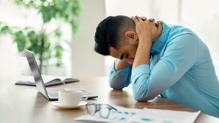 Wall Mural - Young Arab man having strong neck pain or headache, tired of working on laptop at modern office, copy space. Millennial businessman feeling tired, overworking to meet deadline