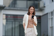 © crizzystudio - Confident businesswoman smiling while using smartphone in front of modern office buildings, showcasing connectivity and mobile technology in business environment