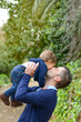 © Nanci - Father lifting playful infant overhead, sharing tender kiss during sunny park moment, green foliage and ivy-covered walls framing affectionate family scene
