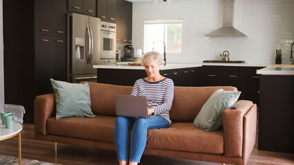 Wall Mural - Senior Woman On Sofa At Home Using Laptop Browsing Online Streaming Show Or Checking Messages