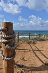 Wall Mural - Rope fence on the beach with blue sky and sea in background in Netanya. Sturdy rope fence, composed of thick, grey ropes secured between light brown wooden posts