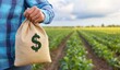 © Emqan - Farmer Holding Money Bag with Dollar Sign in Front of Farmland - Symbol of Financial Success and Sustainable Agriculture