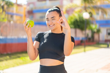 Wall Mural - Young pretty brunette woman with an apple at outdoors showing ok sign with fingers