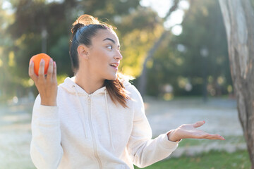 Wall Mural - Young pretty brunette woman holding an orange at outdoors with surprise facial expression