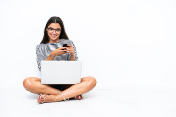 Wall Mural - Young caucasian woman with a laptop sitting on the floor sending a message with the mobile