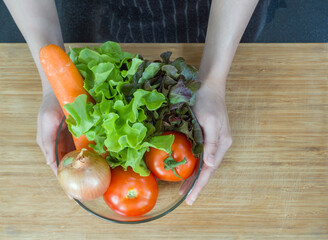 Closeup woman hand holding glass bowl with variety of vegetable on wooden chop board.  Onion, tomato, carrot and lettuce. Raw materials for making  vegetable juice extracts. Top view