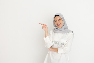 Young muslim woman wearing a grey hijab and white outfit smiles and points to the side while standing against a simple white background presenting something.