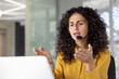 © Liubomir - Serious and focused woman with headset talking remotely, close-up of helpline worker working remotely, using laptop for video call, at workplace inside office.