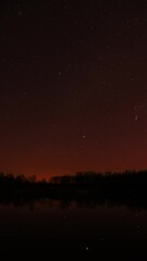 Wall Mural - Real Colorful Night Stars Above Lake River. Natural Starry Sky Background Backdrop. Time Lapse. Vertical Shot. Glowing Stars Reflected In Lake River Water. Natural Starry Sky Background Backdrop