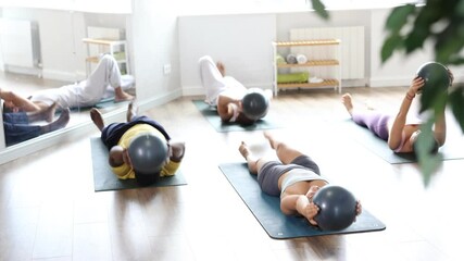 Wall Mural - Diverse group of women and men practicing Pilates in sunlit studio, lying on exercise mats with stability balls in outstretched hands, lifting one leg, focusing on core strength, and coordination