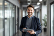 © Pixel Focus AI - Confident young professional dressed smartly, smiling and holding a tablet, standing in a modern office hallway with natural lighting, representing leadership and innovation in a business environment.