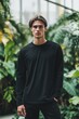 © Aleksander - A young man stands confidently in a lush green environment, wearing a black long sleeve T-shirt mockup. The background features tropical plants, creating a vibrant and natural atmosphere.