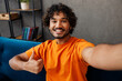 © ViDi Studio - Young man wears casual clothes sits on blue sofa couch do selfie shot pov on mobile cell phone show thumb up stay at home hotel flat rest relax spend free time in living room indoor. Lounge concept.