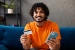 © ViDi Studio - Young man wear casual clothes sit on blue sofa couch use mobile cell phone credit bank card shopping online stay home hotel flat rest relax spend free spare time in living room indoor. Lounge concept