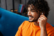 © ViDi Studio - Close up young Indian man wear orange casual clothes sit on blue sofa couch listen to music in headphones stay at home hotel flat rest relax spend free spare time in living room indoor Lounge concept
