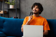 © ViDi Studio - Young Indian man wear orange casual clothes sits on blue sofa couch use laptop pc computer hold credit bank card shopping online stay home hotel flat rest relax spend free time in living room indoor.
