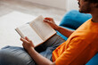 © ViDi Studio - Close up side view young Indian man wears orange casual clothes sits on blue sofa couch read novel book stay at home hotel flat rest relax spend free spare time in living room indoor. Lounge concept.
