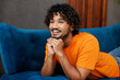 © ViDi Studio - Side profile view young smiling happy Indian man wear orange casual clothes lay down on blue sofa couch stay at home hotel flat rest relax spend free spare time in living room indoor. Lounge concept.