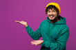 © ViDi Studio - Young cheerful happy Indian man he wear green hoody yellow hat casual clothes point hands arms aside on area mockup look camera isolated on plain purple background studio portrait. Lifestyle concept.