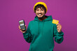 © ViDi Studio - Young happy Indian man he wear green hoody yellow hat casual clothes hold wireless modern bank payment terminal to process acquire credit card isolated on plain purple background. Lifestyle concept.