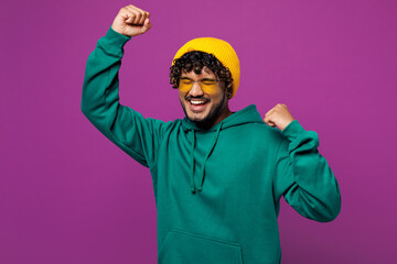 Wall Mural - Young overjoyed excited happy cool Indian man he wear green hoody yellow hat sunglasses casual clothes do winner gesture dance isolated on plain purple background studio portrait. Lifestyle concept.