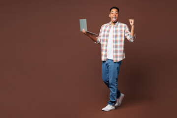 Wall Mural - Full body happy young IT man of African American ethnicity he wear blue shirt casual clothes hold use work on laptop pc computer isolated on plain brown background studio portrait. Lifestyle concept.