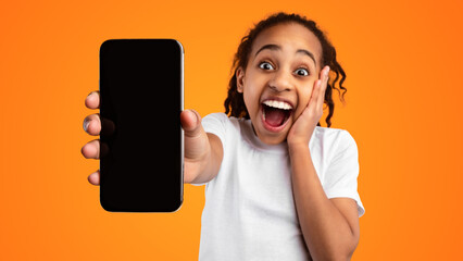 Wow, Amazing Offer. Portrait of excited African American girl holding mobile phone with empty screen in hand, showing device close to camera and touching cheek, mock up, yellow studio background
