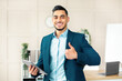 © Prostock-studio - Smiling Arab businessman holding computer, showing thumb up gesture in office. Handsome guy entrepreneur recommending online services, having virtual conference with colleagues