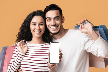 Poster - Online Shopping App. Cheerful young arab couple showing smartphone with blank white screen at camera and holding bright shopper bags while standing isolated over yellow studio background, mockup