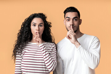 Wall Mural - Young arab man and woman showing shh gesture at camera, mysterious middle eastern couple keeping forefingers on lips, making silence sign while standing together over yellow background, copy space