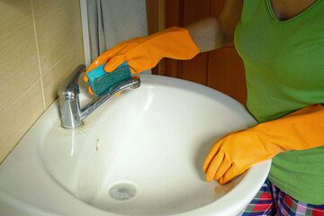 Woman in pajamas doing housework: Close-up of female hands in orange gloves washing dirty ceramic sink and faucet in bathroom with sponge. Saving on services: do-it-yourself house cleaning.