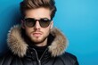 © Bay - A fashionable man showcases a youthful winter outfit with a fur-collared jacket and stylish sunglasses, standing confidently in front of a bright blue background