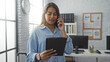 © Krakenimages.com - Woman talking on the phone while holding a tablet in a modern office workspace with large windows and white brick walls in an indoor setting.