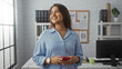 © Krakenimages.com - Woman smiling indoors in an office setting, holding a smartphone while wearing a light blue shirt, surrounded by shelved books, plants, and a window with natural light.